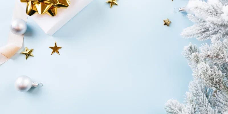 White gift box with gold bow and silver ornaments arranged on pale blue background with snowy pine branches and scattered stars