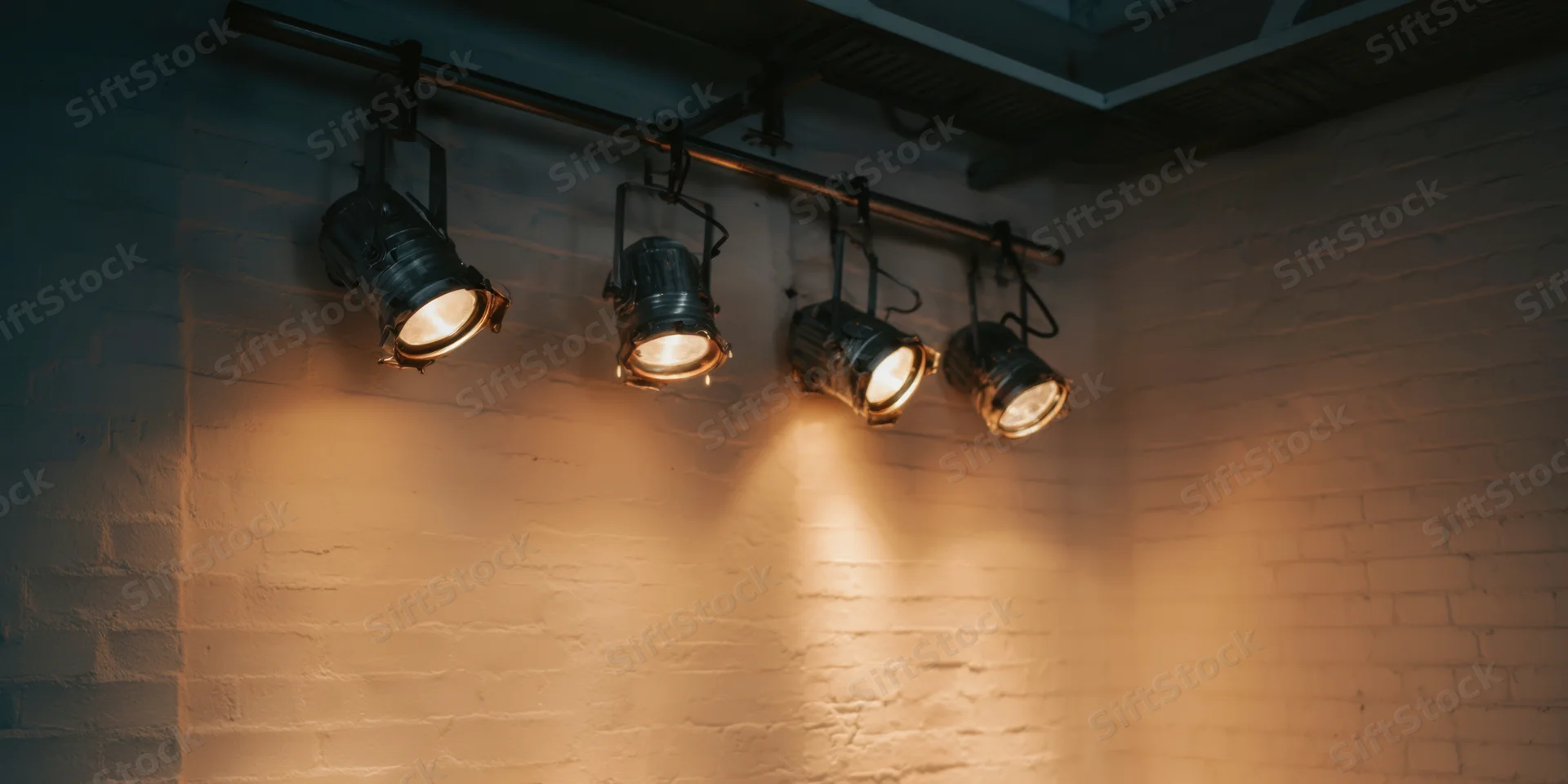 Four industrial spotlights hanging in a row on a track above a white brick wall, casting warm beams of light