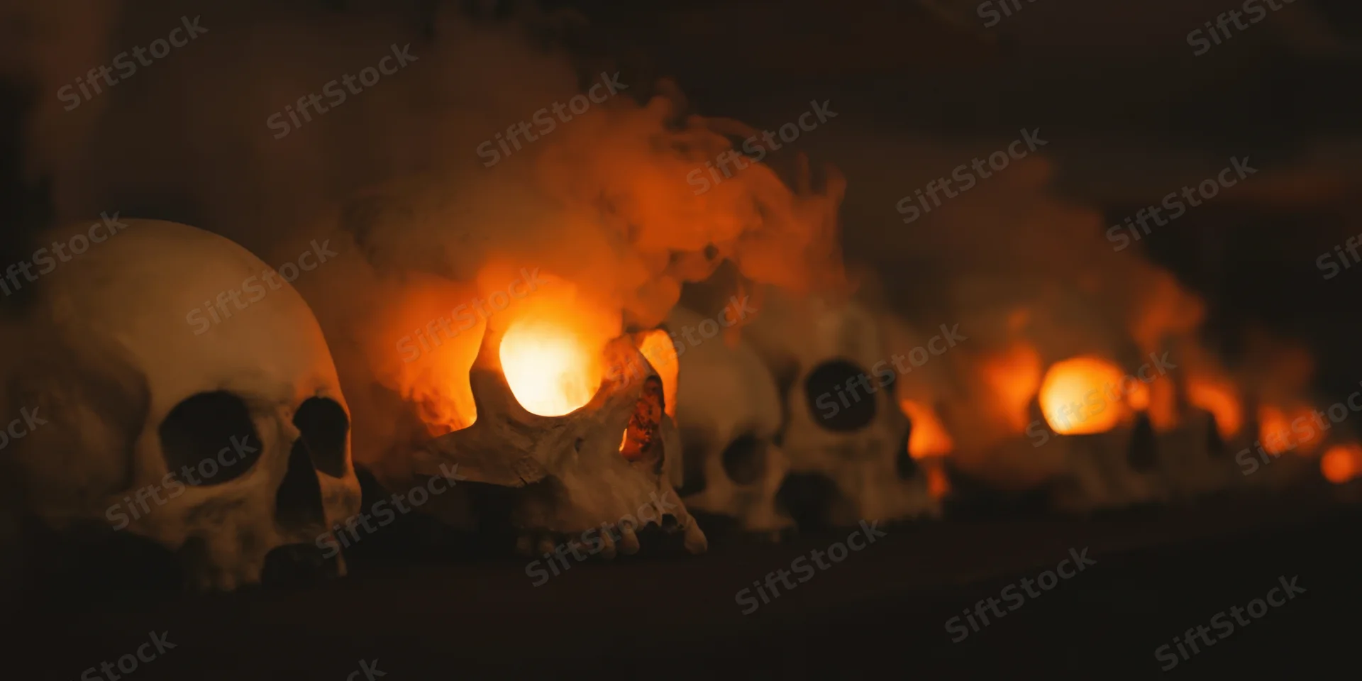 Row of skulls arranged in smoky dimly lit scene with orange flames glowing from within
