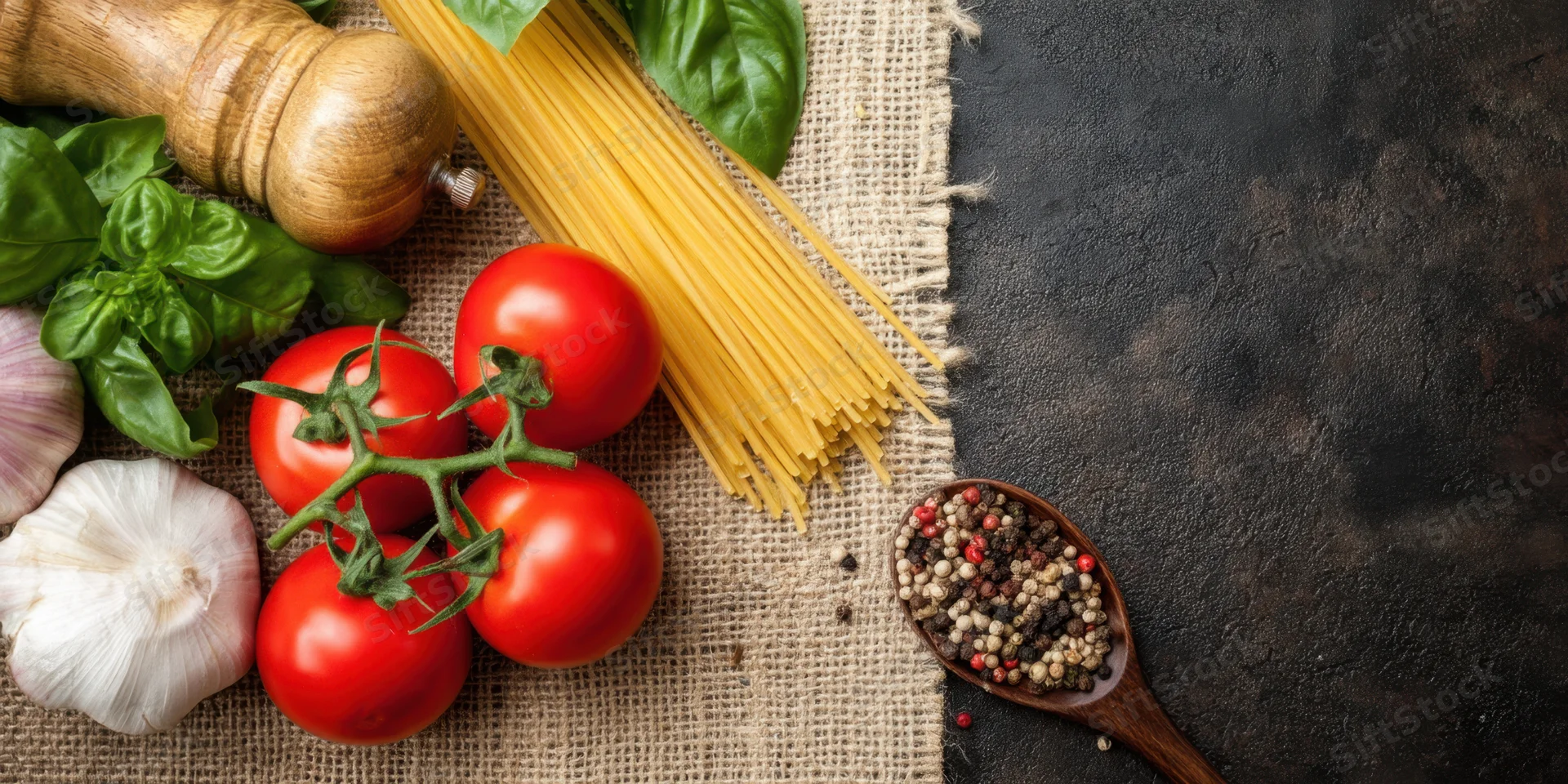 Tomatoes on the vine with spaghetti, basil, garlic, pepper and wooden spoon arranged on burlap and dark surface