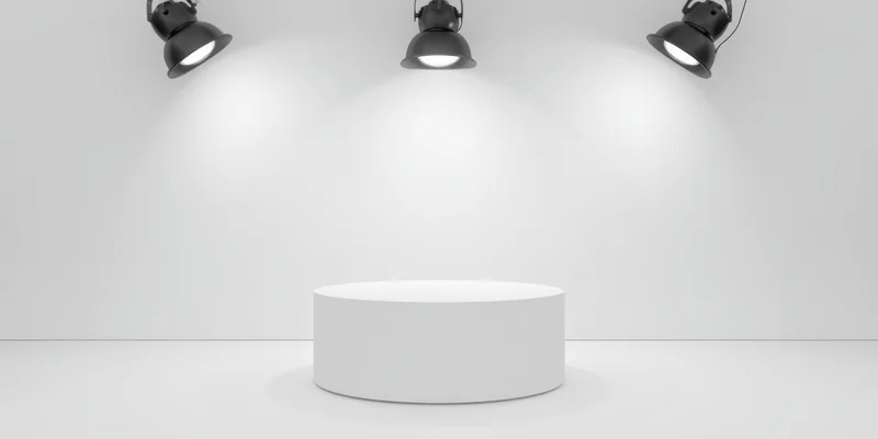 White cylindrical pedestal under three overhead spotlights in a minimalist studio space