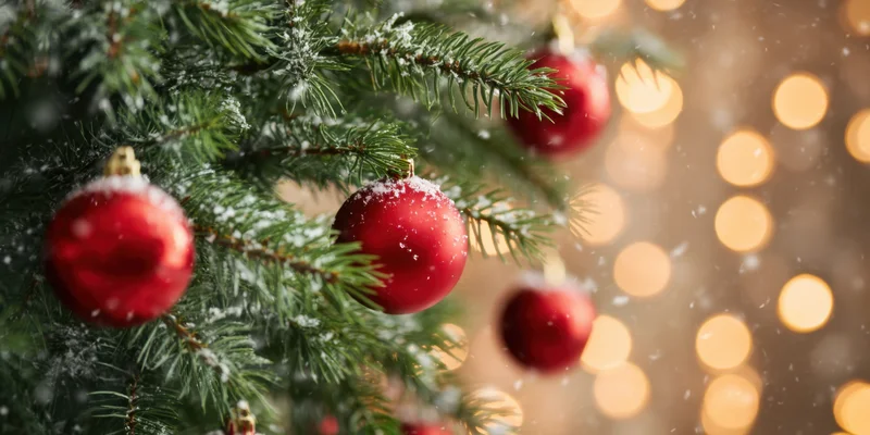 Close up of evergreen christmas tree branches with red baubles and snow, warm bokeh lights in background