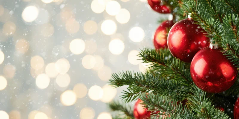 Close up of evergreen christmas tree branches with shiny red ornaments against blurred bokeh lights and falling snow