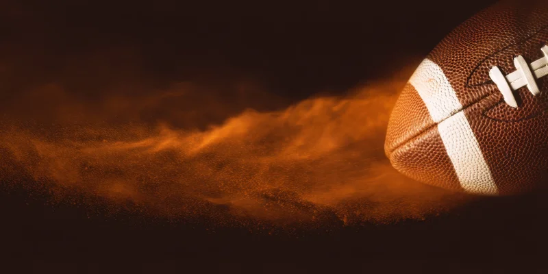 Close-up of an american football with textured brown leather and white laces, surrounded by glowing orange dust in motion against a dark background