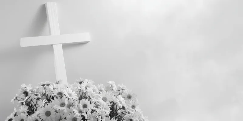 White cross leaning against pale wall with bouquet of white daisies at base