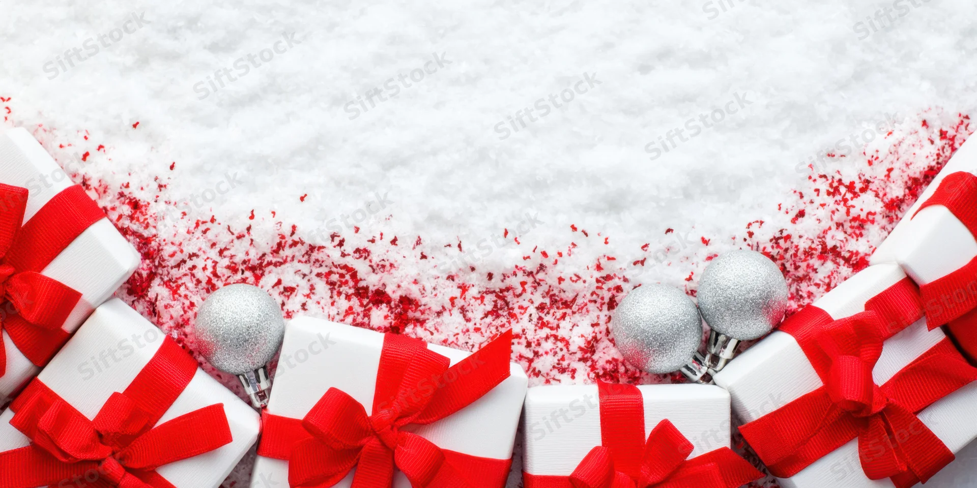 White gift boxes with red ribbons arranged in a row on snowy surface with silver ornaments and red confetti