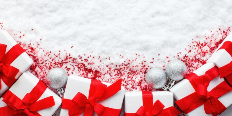 White gift boxes with red ribbons arranged in a row on snowy surface with silver ornaments and red confetti