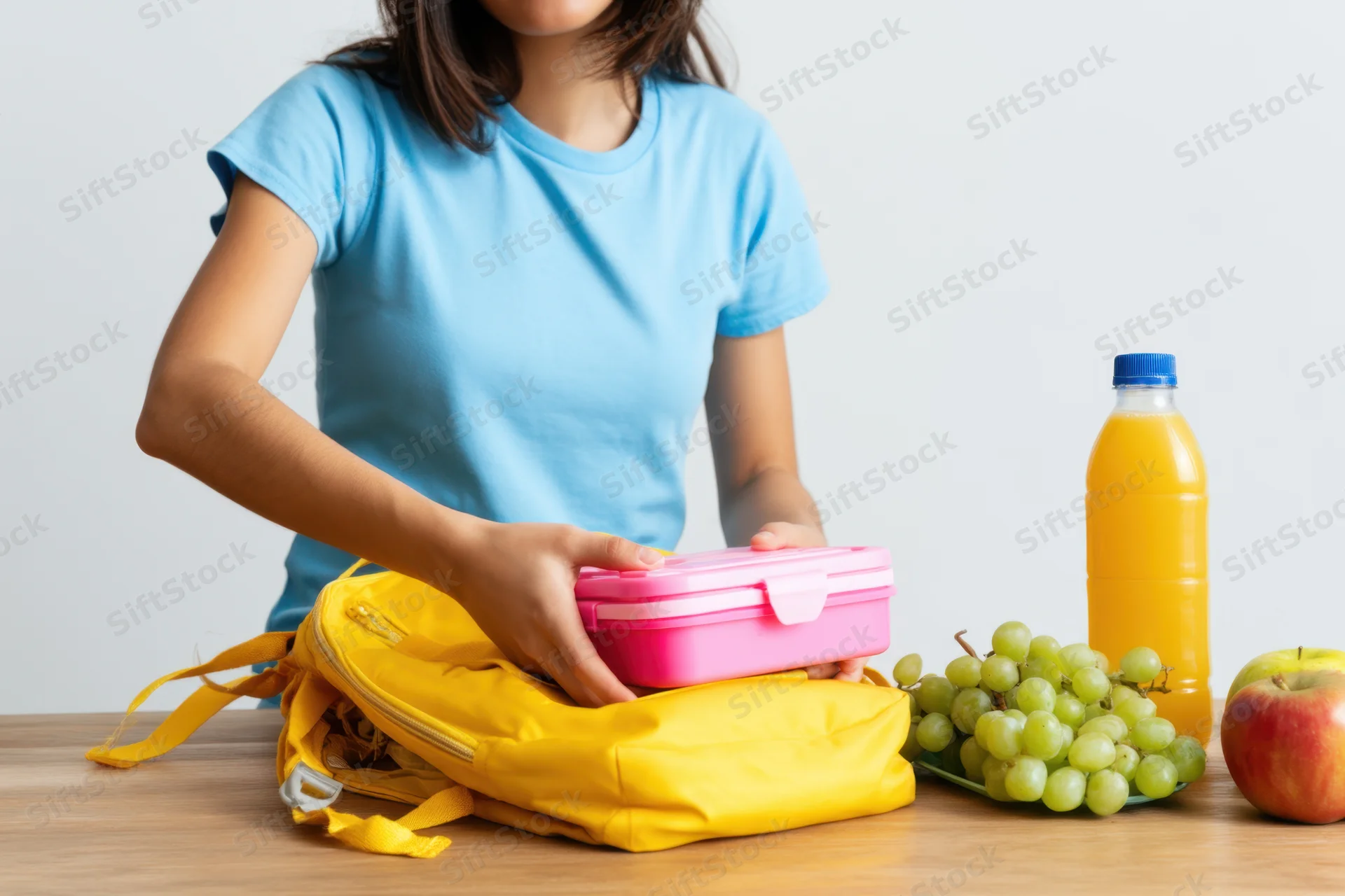 Child placing pink lunchbox into yellow backpack on wooden table with fruit and juice nearby