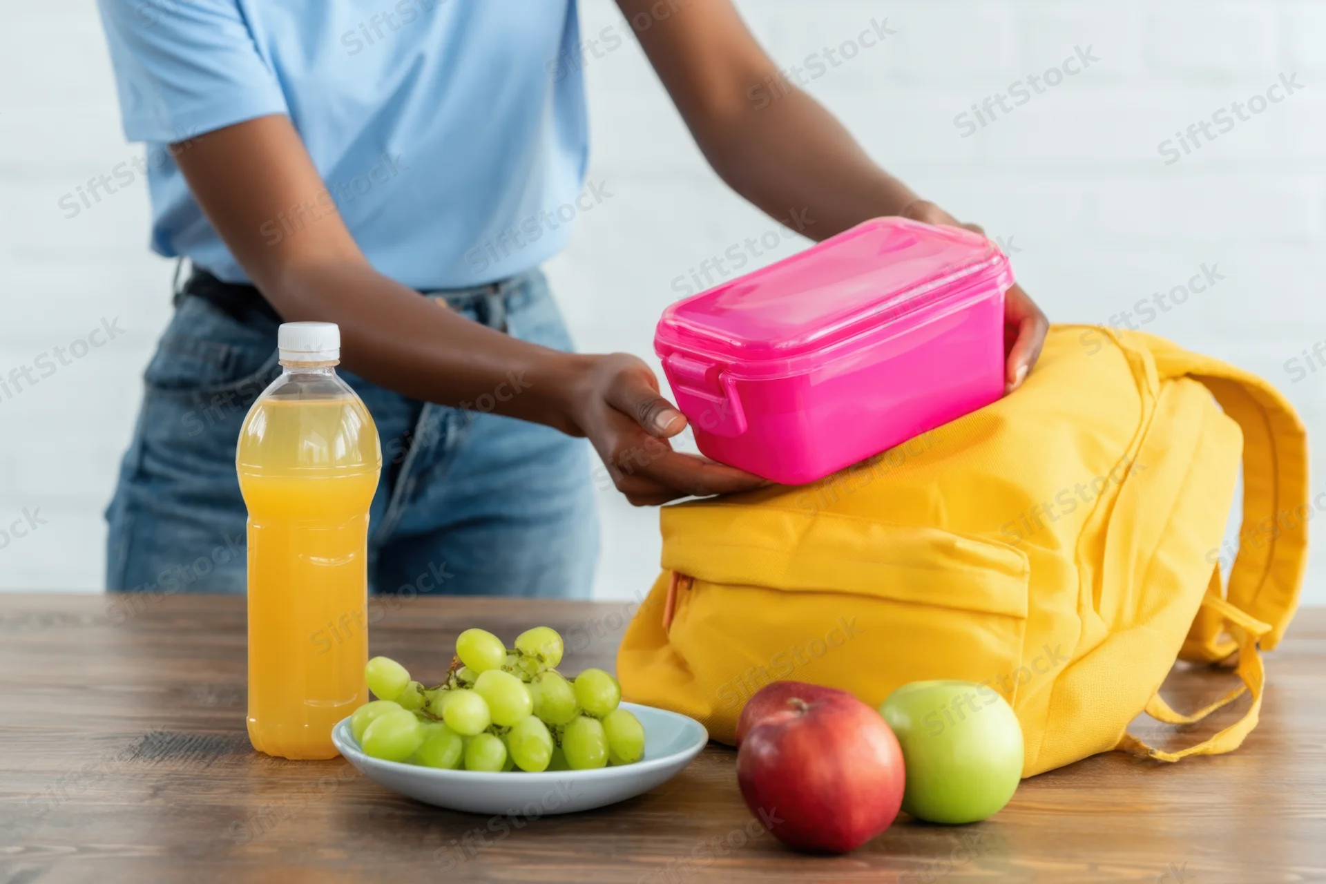 Student placing pink lunchbox into yellow backpack on wooden table with fruit and juice nearby