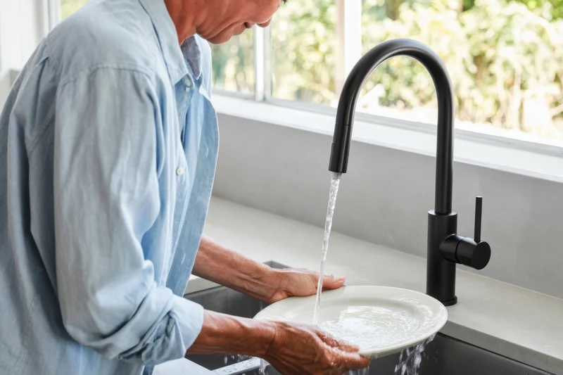 Man washing a white plate under running water at a kitchen sink near a window