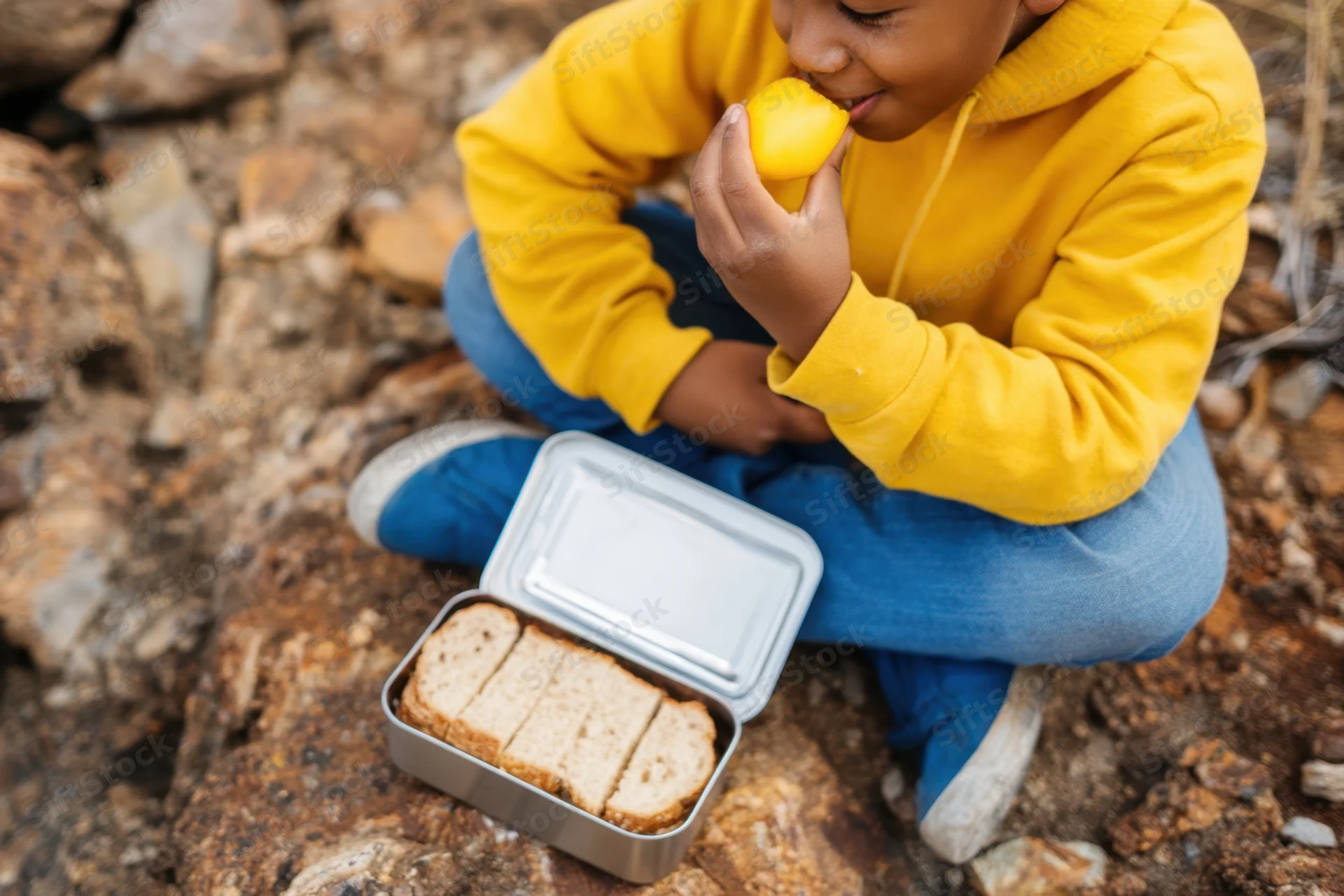 Child eating orange and sandwich from lunchbox while sitting on rocky ground outdoors