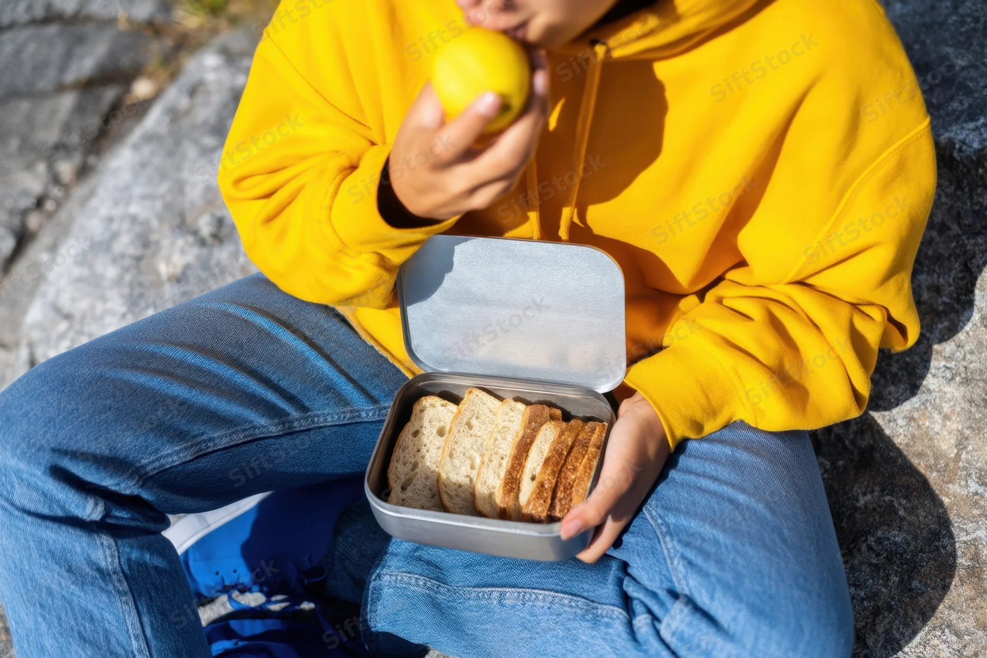 Person in yellow hoodie eating lemon and holding sandwich container while sitting on rocky ground outdoors