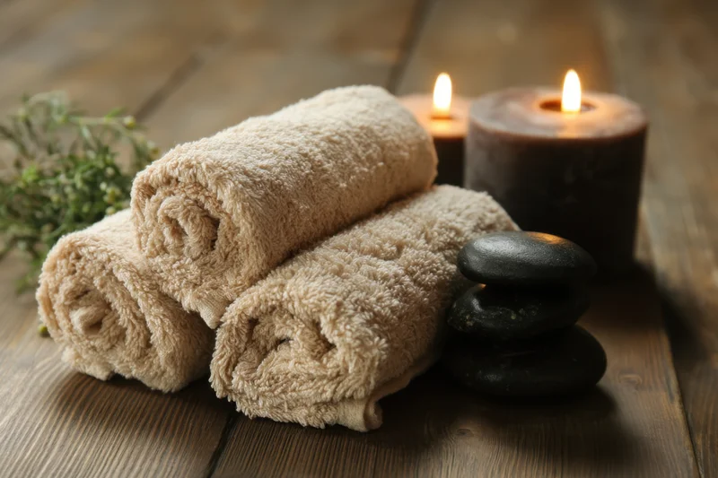 Rolled beige towels stacked beside lit candles and smooth black stones on a wooden table