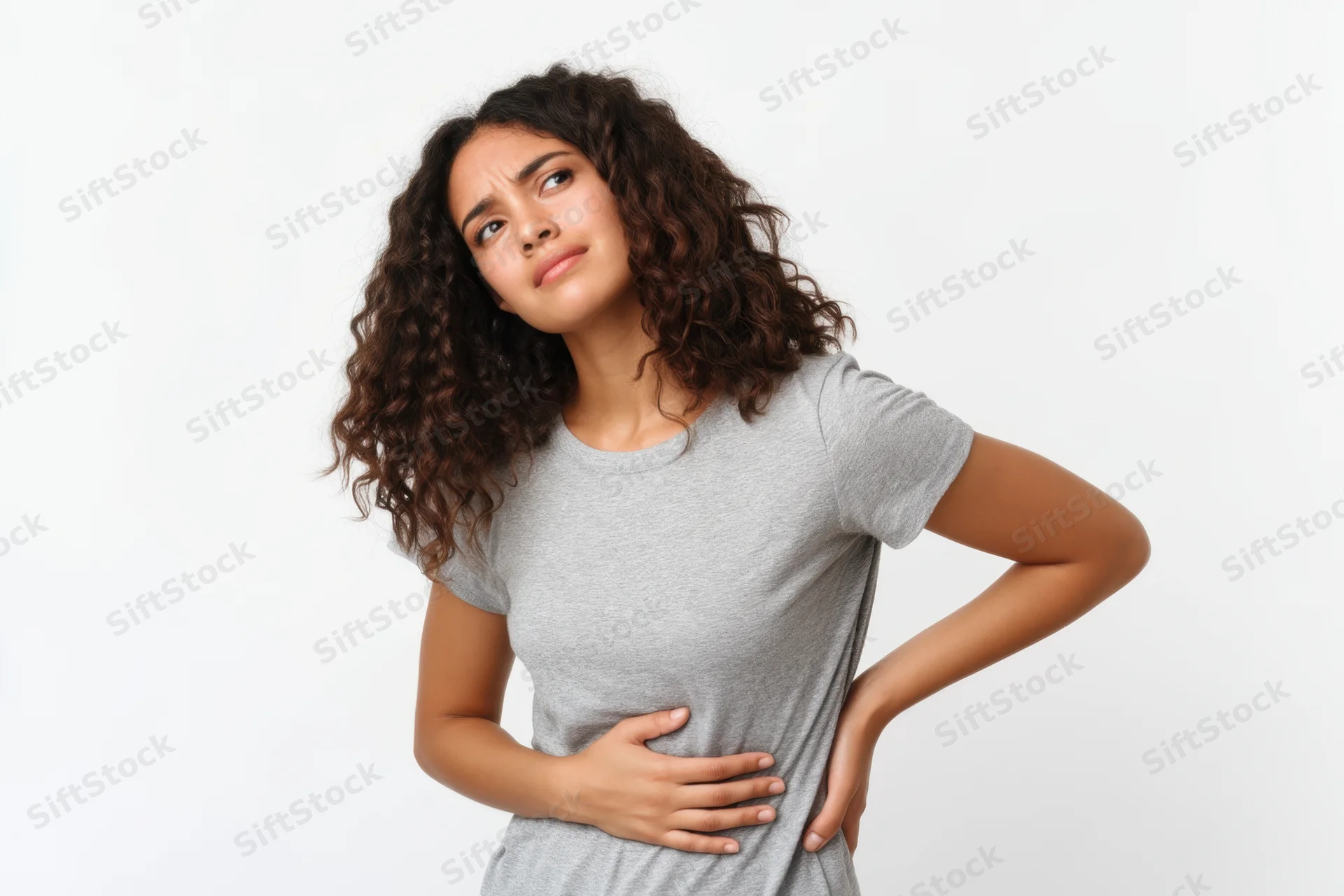 Young hispanic woman in gray t-shirt standing with hands on stomach showing discomfort against white studio background
