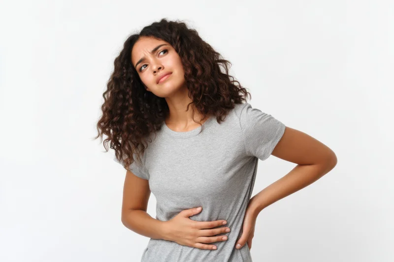 Young hispanic woman in gray t-shirt standing with hands on stomach showing discomfort against white studio background