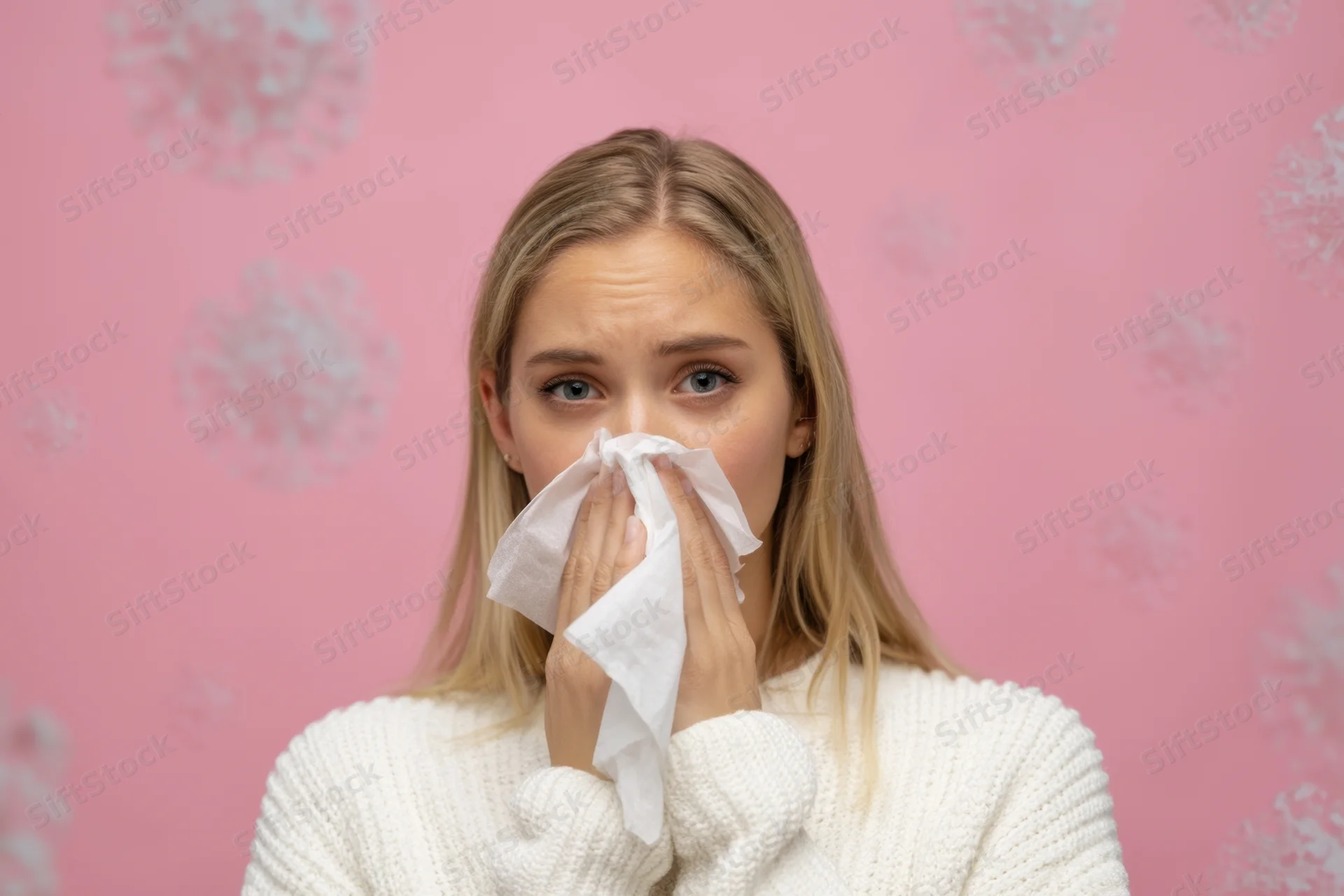 Young woman blowing her nose with a tissue against a pink patterned background