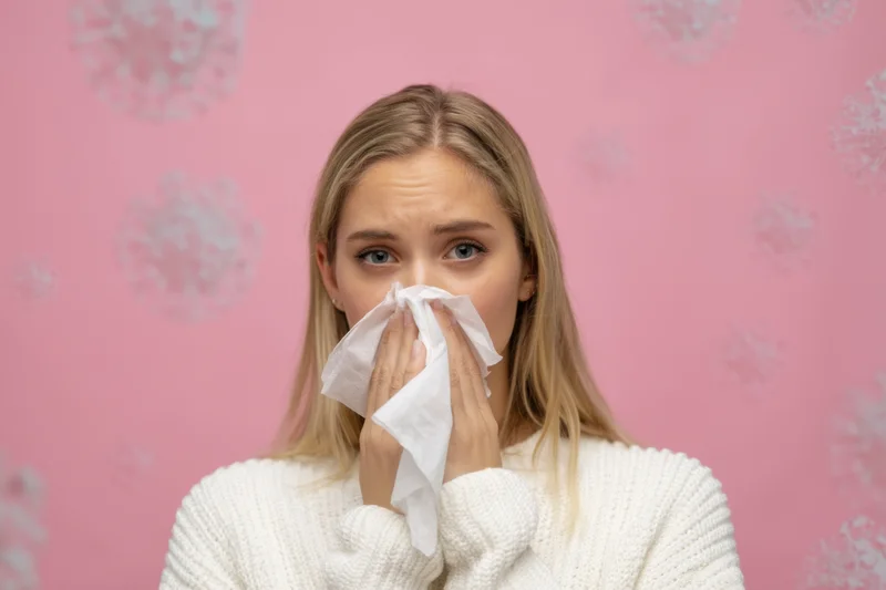 Young woman blowing her nose with a tissue against a pink patterned background