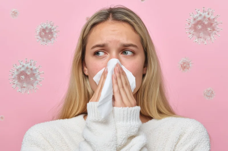 Young woman blowing her nose with tissue against pink background showing virus particles