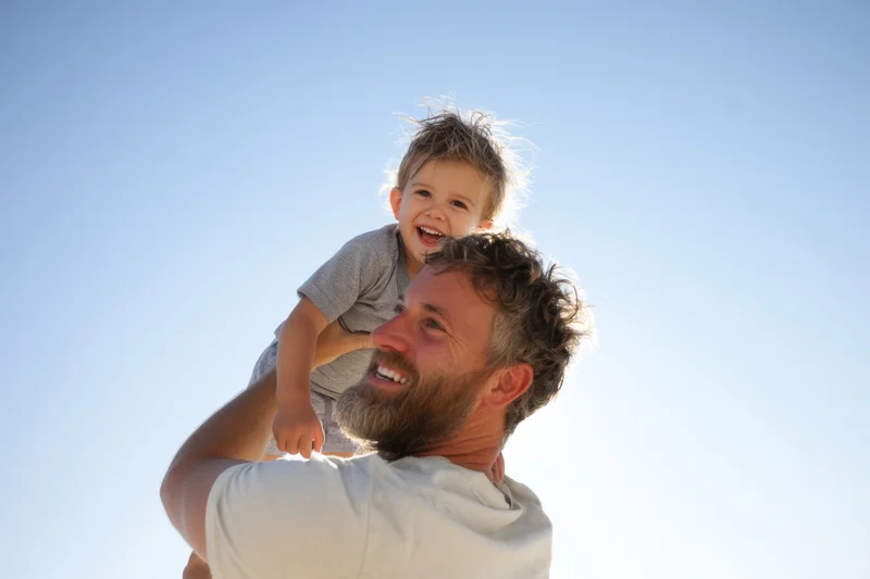 Father carrying laughing child on shoulders against clear blue sky
