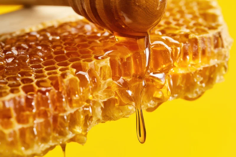 Honey dripping from a wooden dipper onto a honeycomb structure against a yellow background