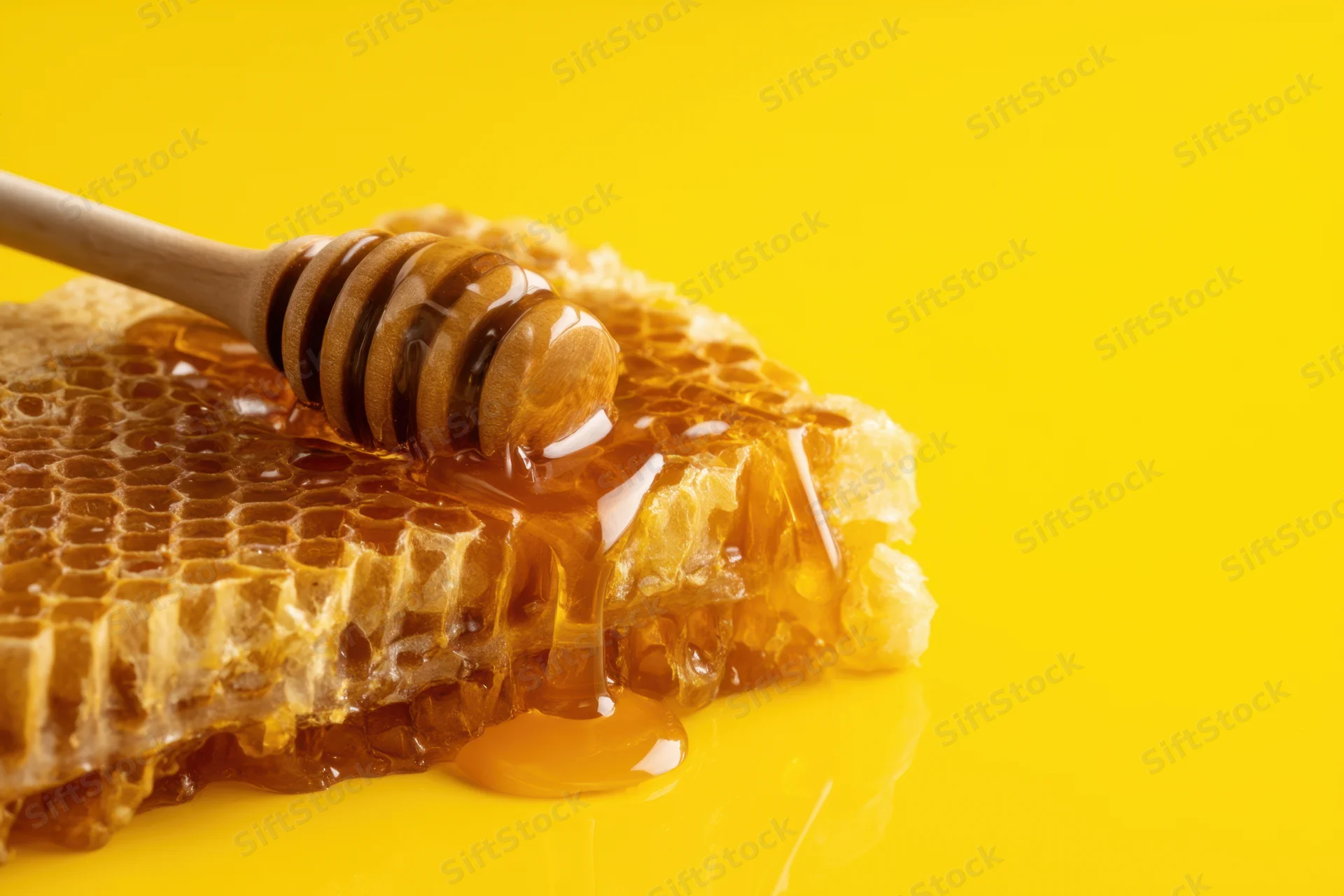 Honey dripping from a wooden dipper onto a honeycomb structure on a yellow surface
