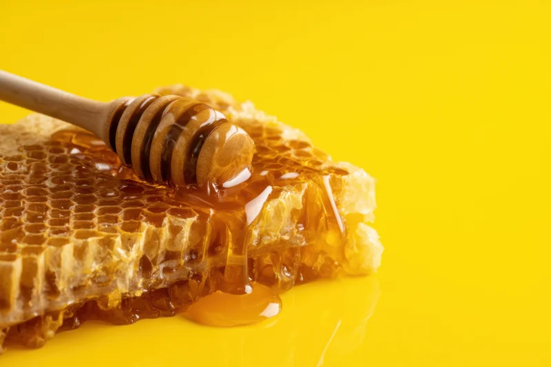 Honey dripping from a wooden dipper onto a honeycomb structure on a yellow surface