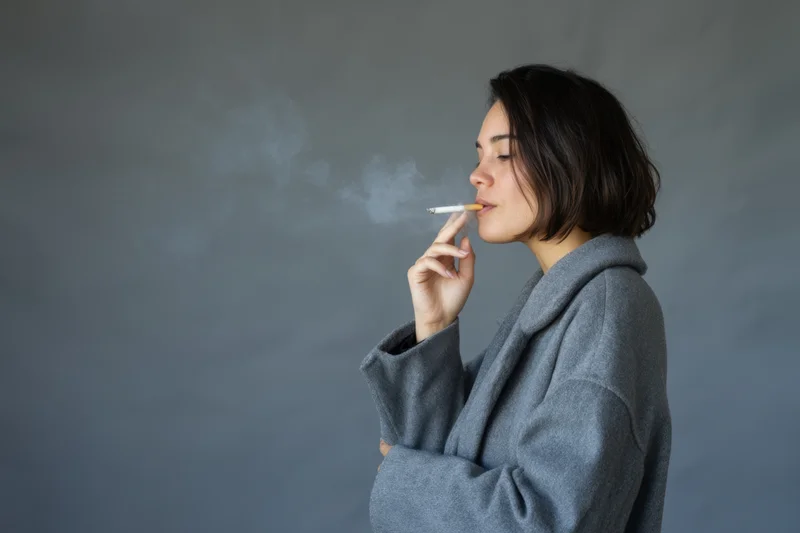 Woman smoking a cigarette in profile in a gray coat against a plain background