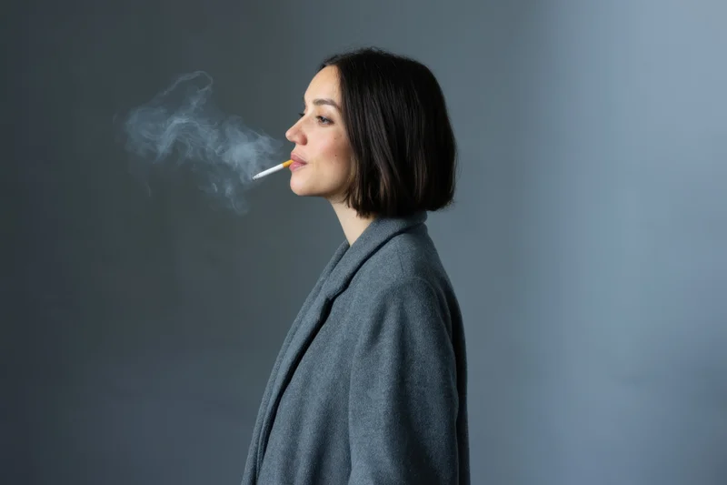 Woman in gray coat smoking cigarette in studio profile view exhaling smoke
