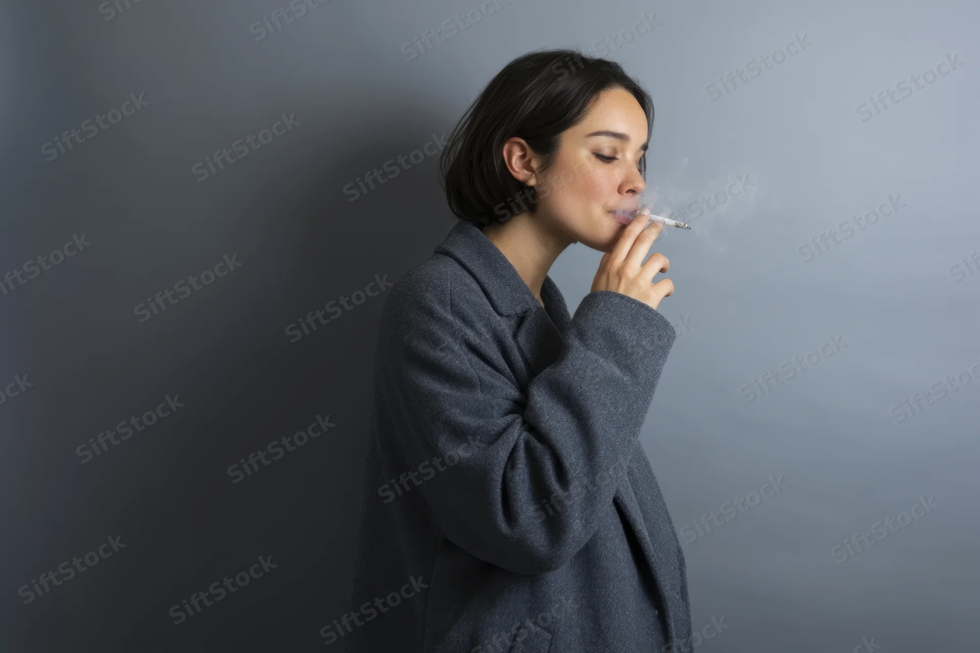 Woman smoking cigarette in studio portrait, exhaling smoke, side profile, gray coat, neutral background