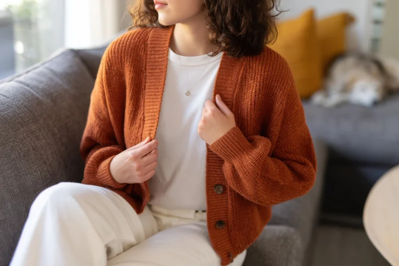 Woman seated on gray fabric sofa wearing rust cardigan over white tshirt and pants, hand on chest, cat resting on cushion behind