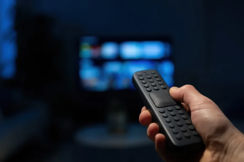 Hand holding remote control aimed at blurred television screen in dimly lit room