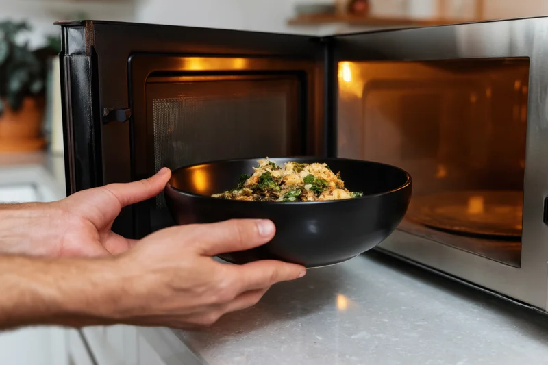 Hands removing a black bowl of reheated food from an open microwave on a kitchen countertop