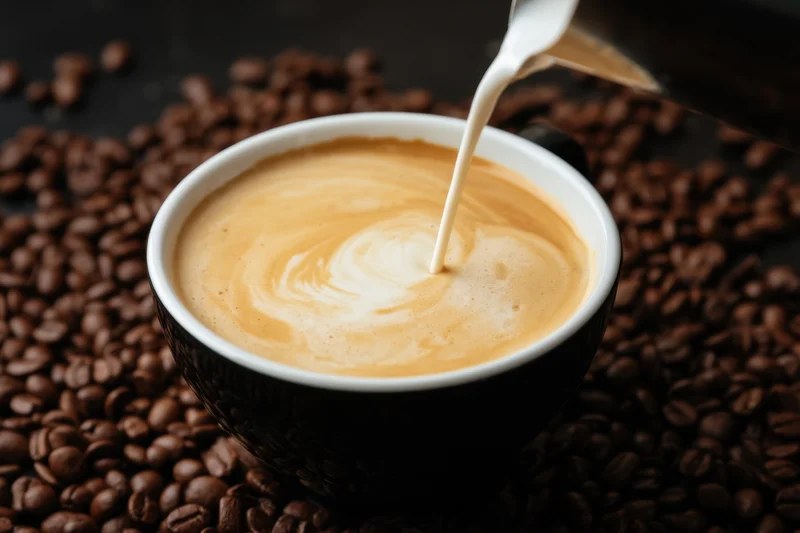 Milk being poured into a black coffee cup with latte art forming on top surrounded by coffee beans