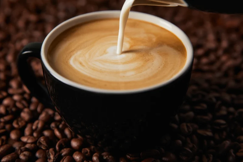 Milk being poured into a black coffee cup creating latte art on a bed of coffee beans