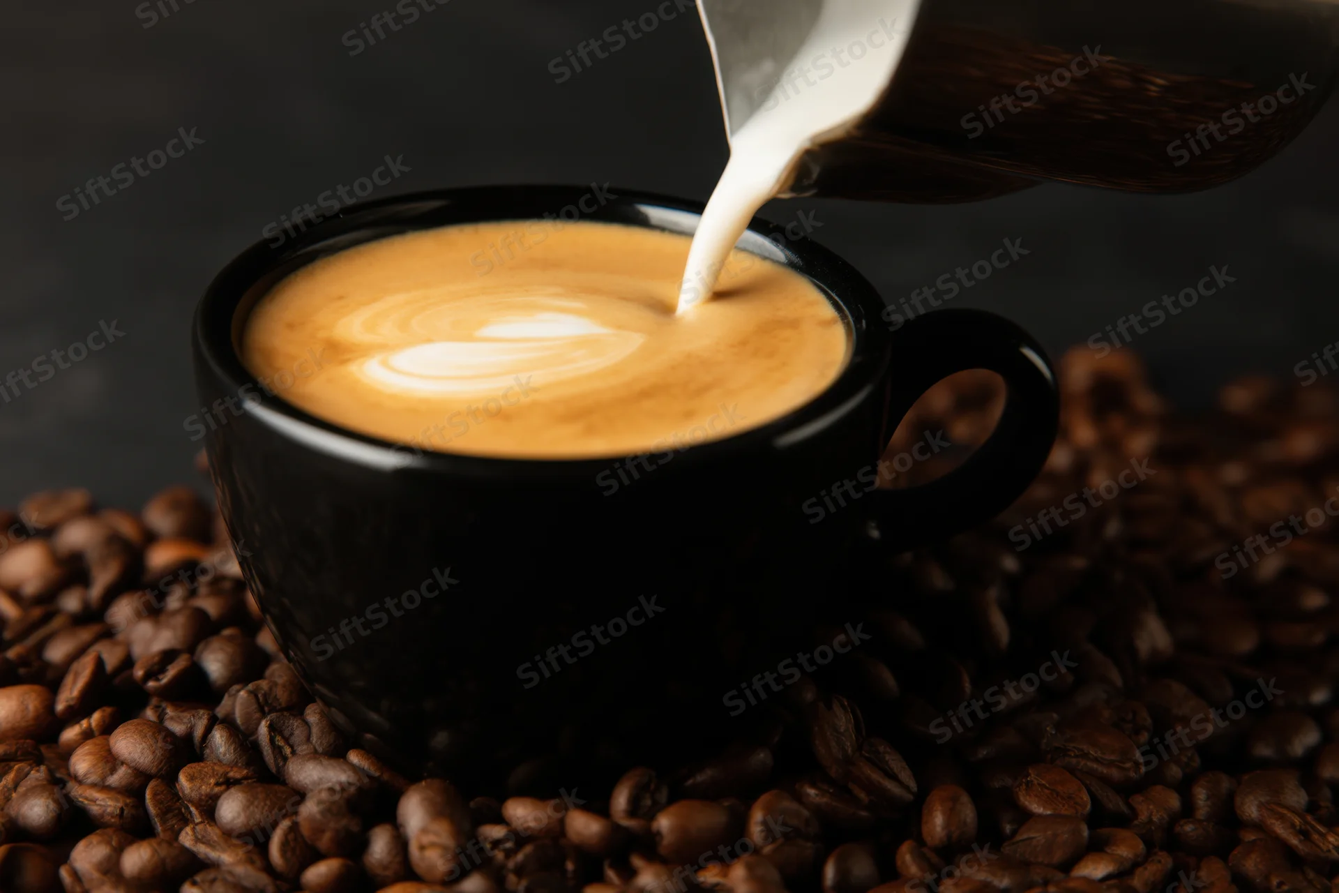 Milk being poured into a black coffee cup with latte art on a bed of coffee beans