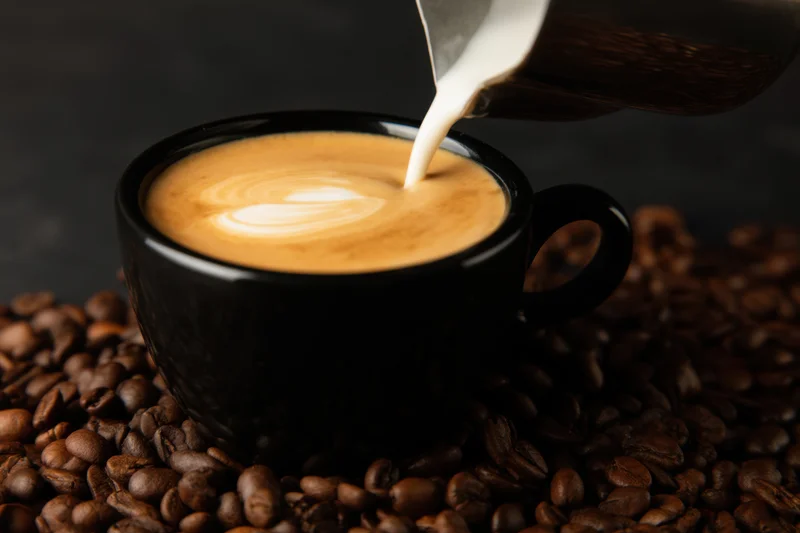 Milk being poured into a black coffee cup with latte art on a bed of coffee beans