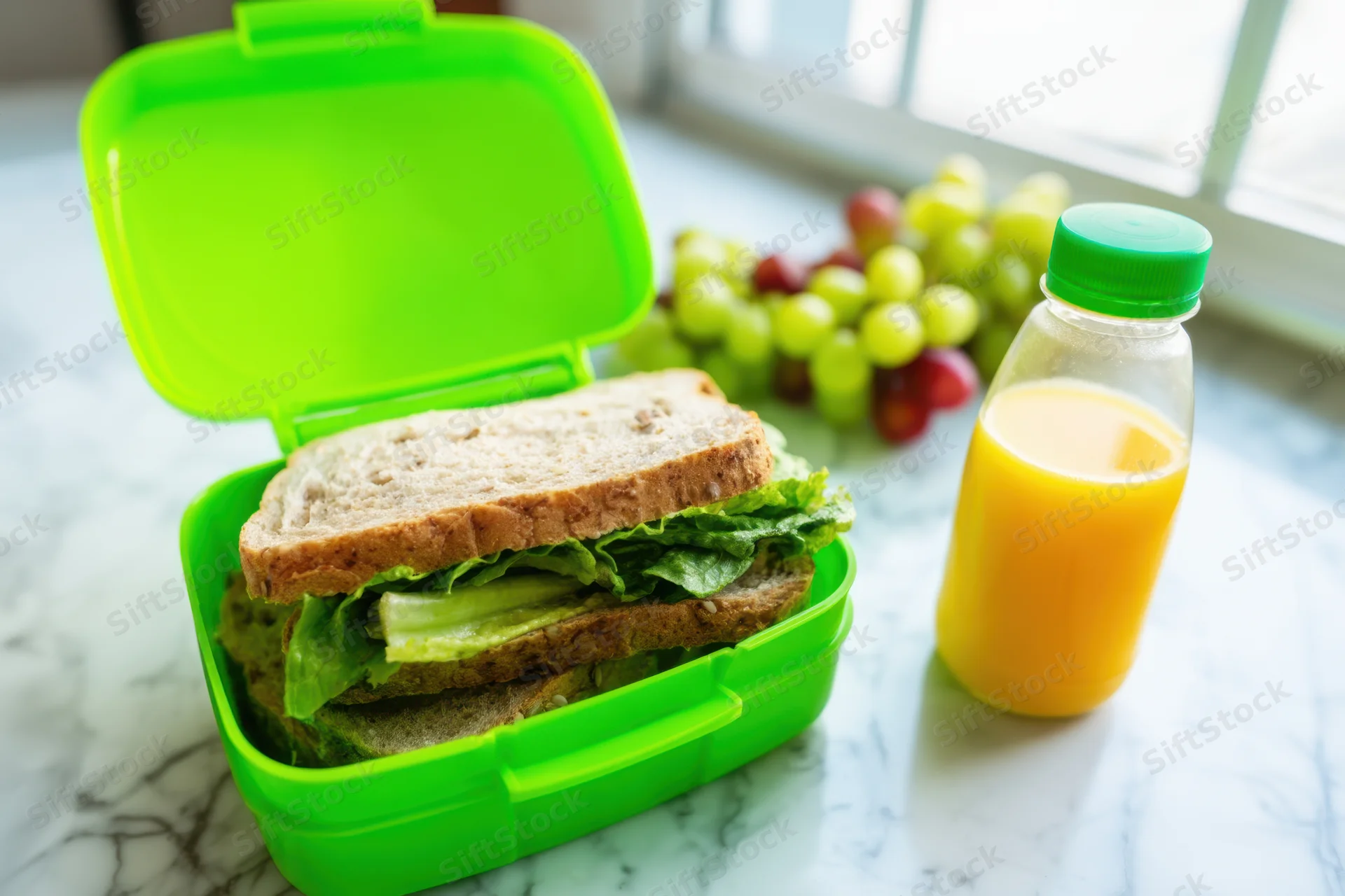 Open green lunchbox with sandwich and grapes on marble countertop near window