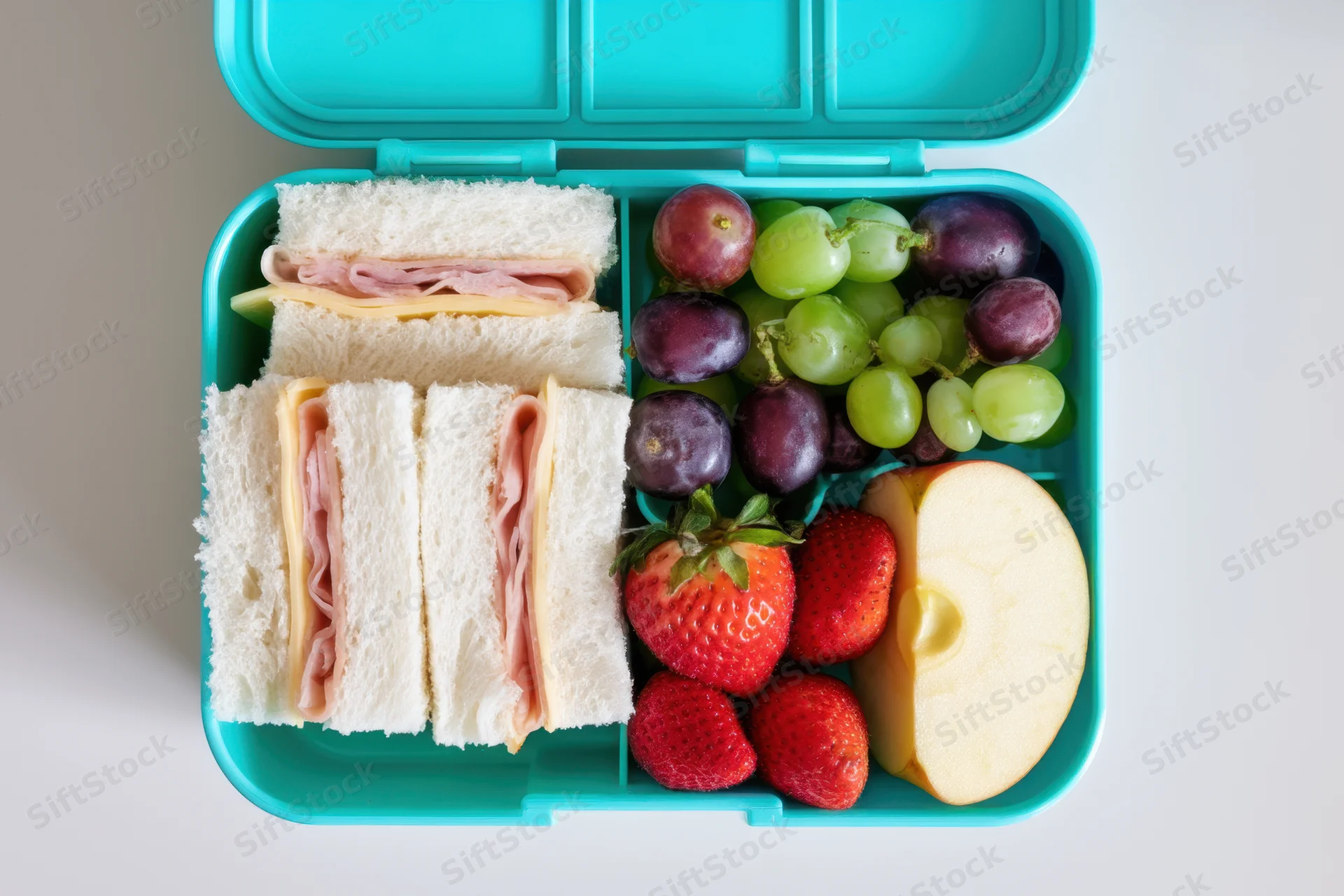 Turquoise lunchbox with sandwiches and fruit arranged in compartments on a white surface