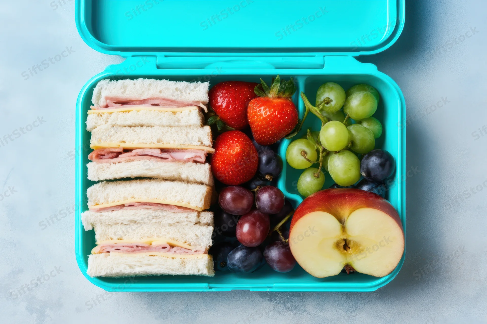Turquoise lunchbox with sandwiches and assorted fruits arranged in compartments on a light surface