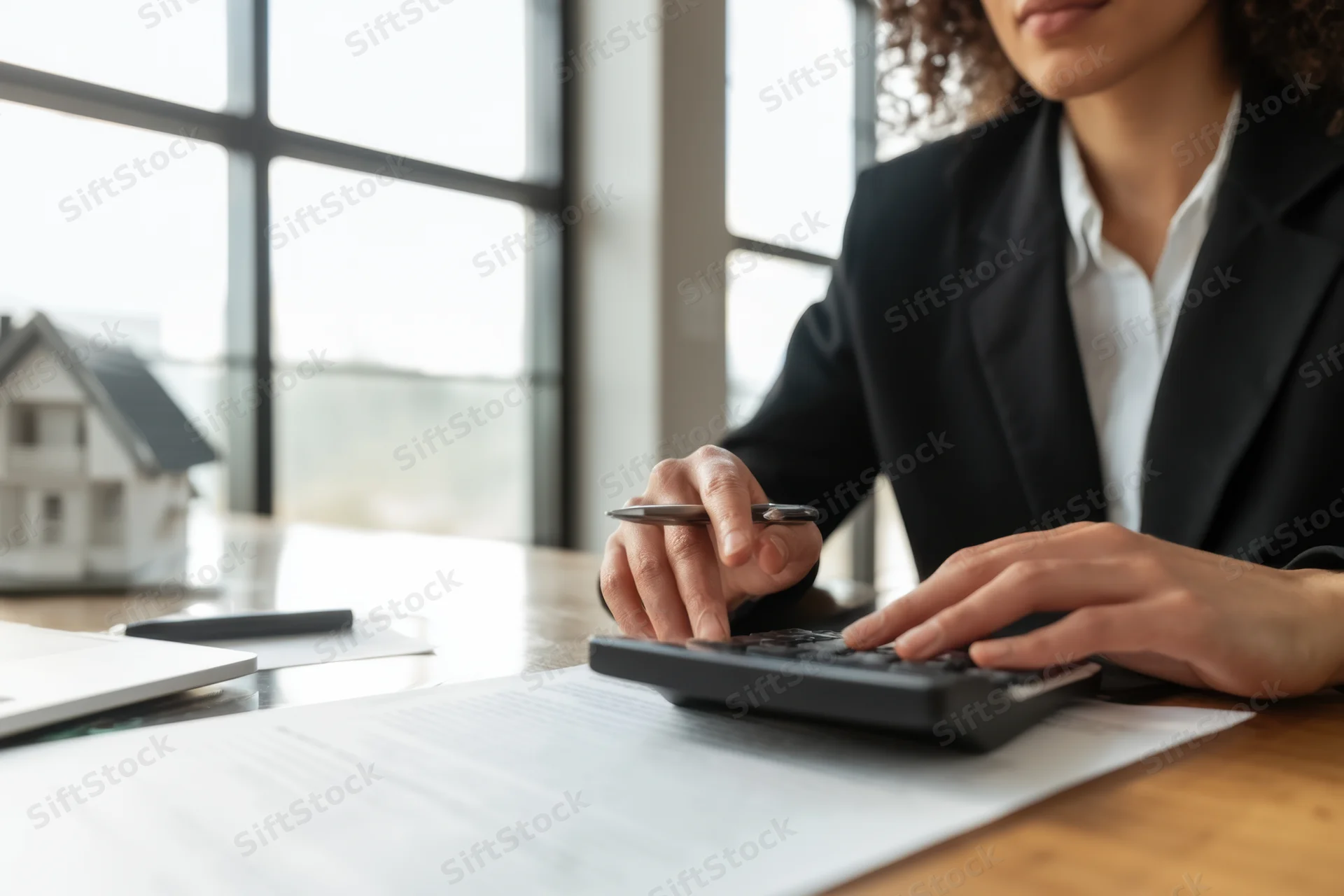 Woman in black blazer using calculator on desk with house model and documents in office
