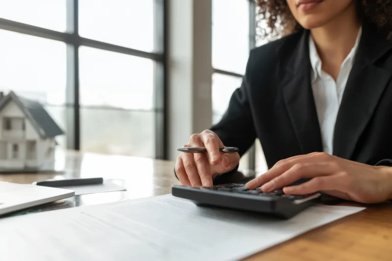 Woman in black blazer using calculator on desk with house model and documents in office
