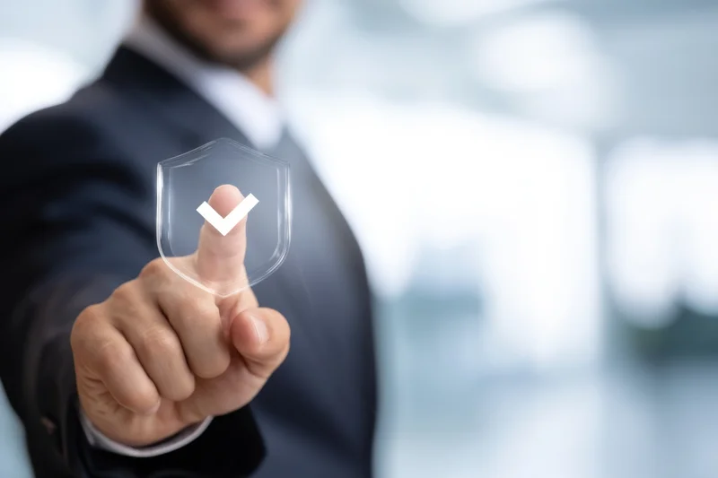 Businessman in dark suit pointing at transparent digital shield with checkmark icon in office setting