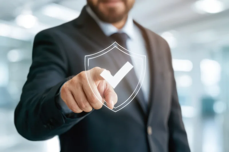 Businessman in dark suit pointing at transparent digital shield with checkmark in office interior