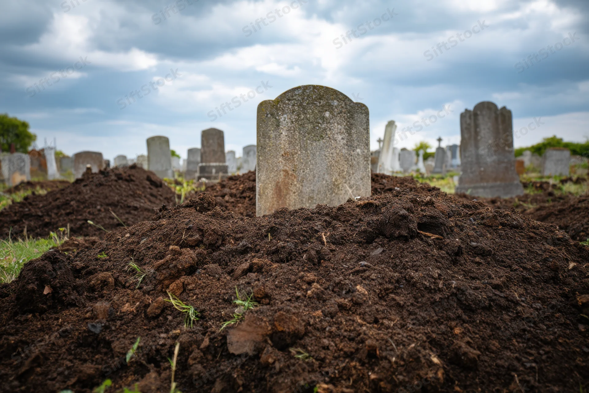 Freshly dug grave mound with weathered tombstone in cemetery under cloudy sky
