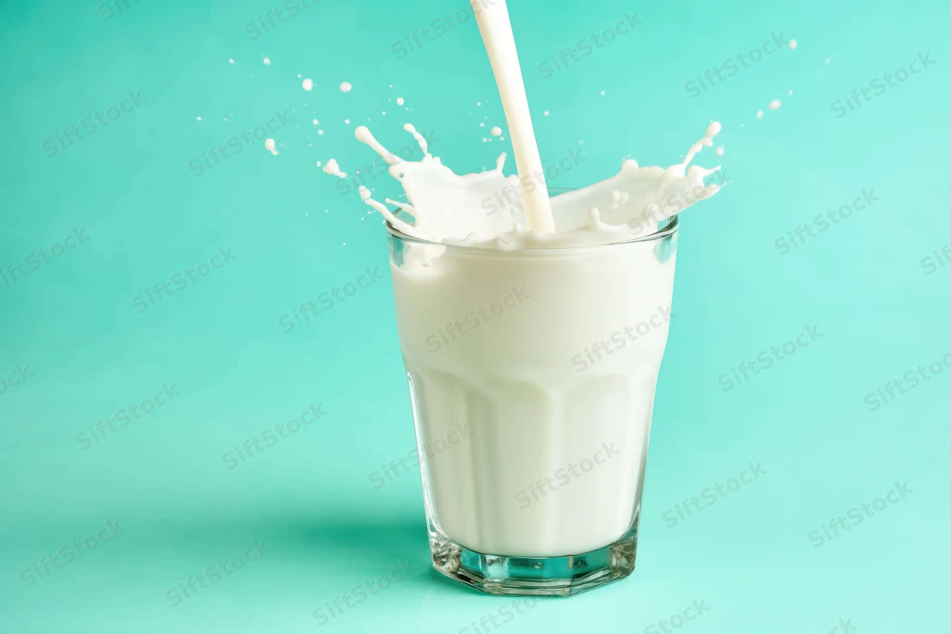 Milk being poured into a clear glass with splash against teal background