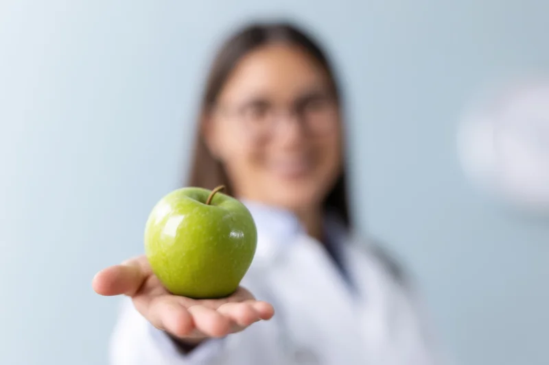 Woman in lab coat holding green apple in hand, smiling, with blurred background
