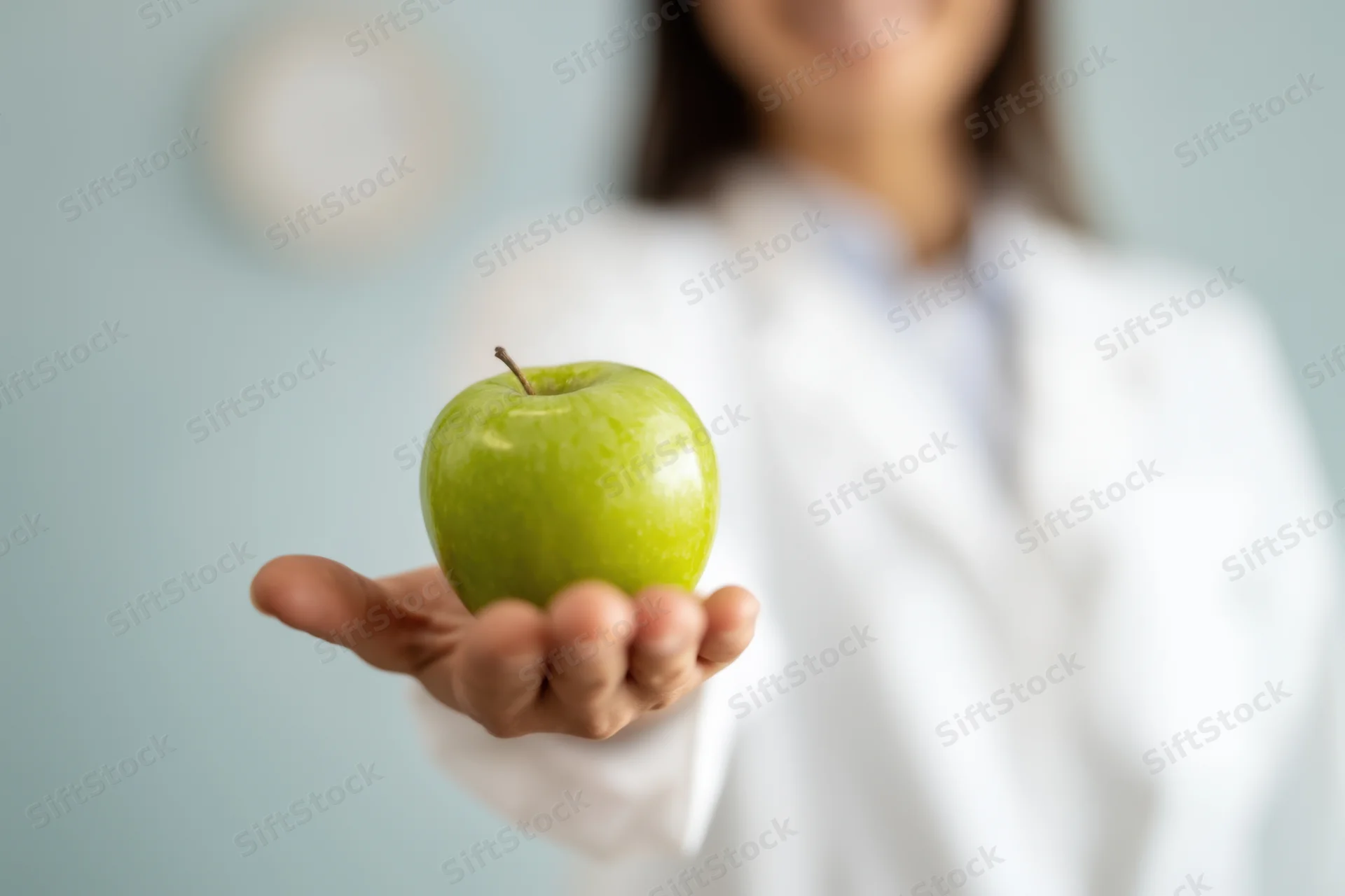 Woman in white coat holding green apple in hand, close-up view with blurred background