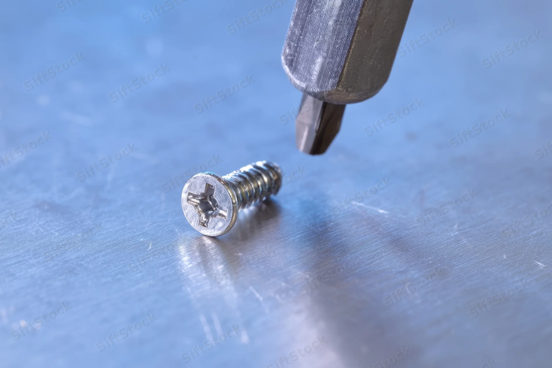 Metallic screw lying on a brushed metal surface with a screwdriver bit hovering above it
