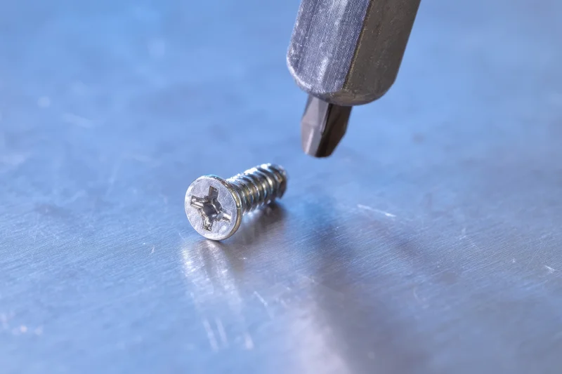 Metallic screw lying on a brushed metal surface with a screwdriver bit hovering above it