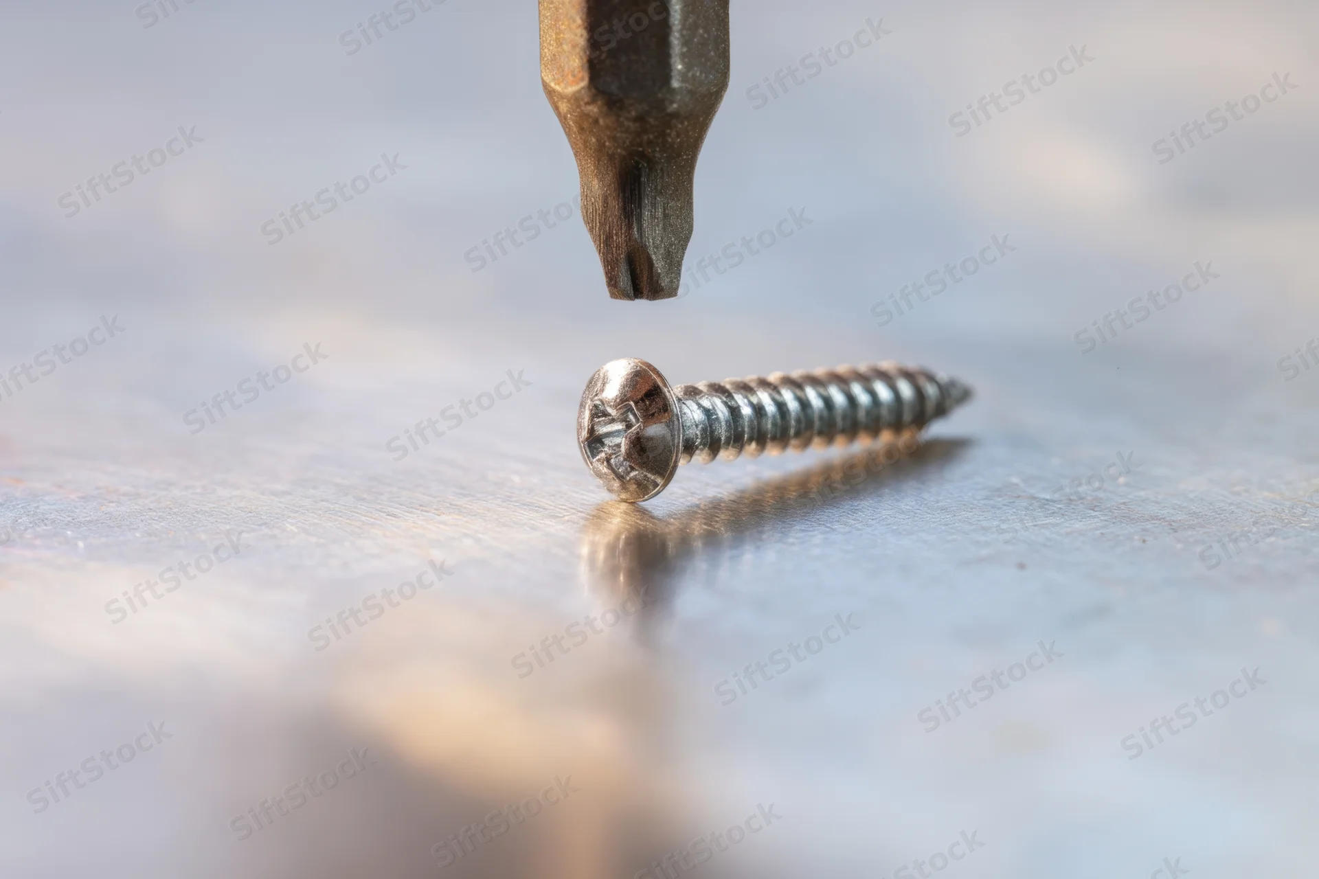 Metallic screw lying on wooden surface with screwdriver bit hovering above it